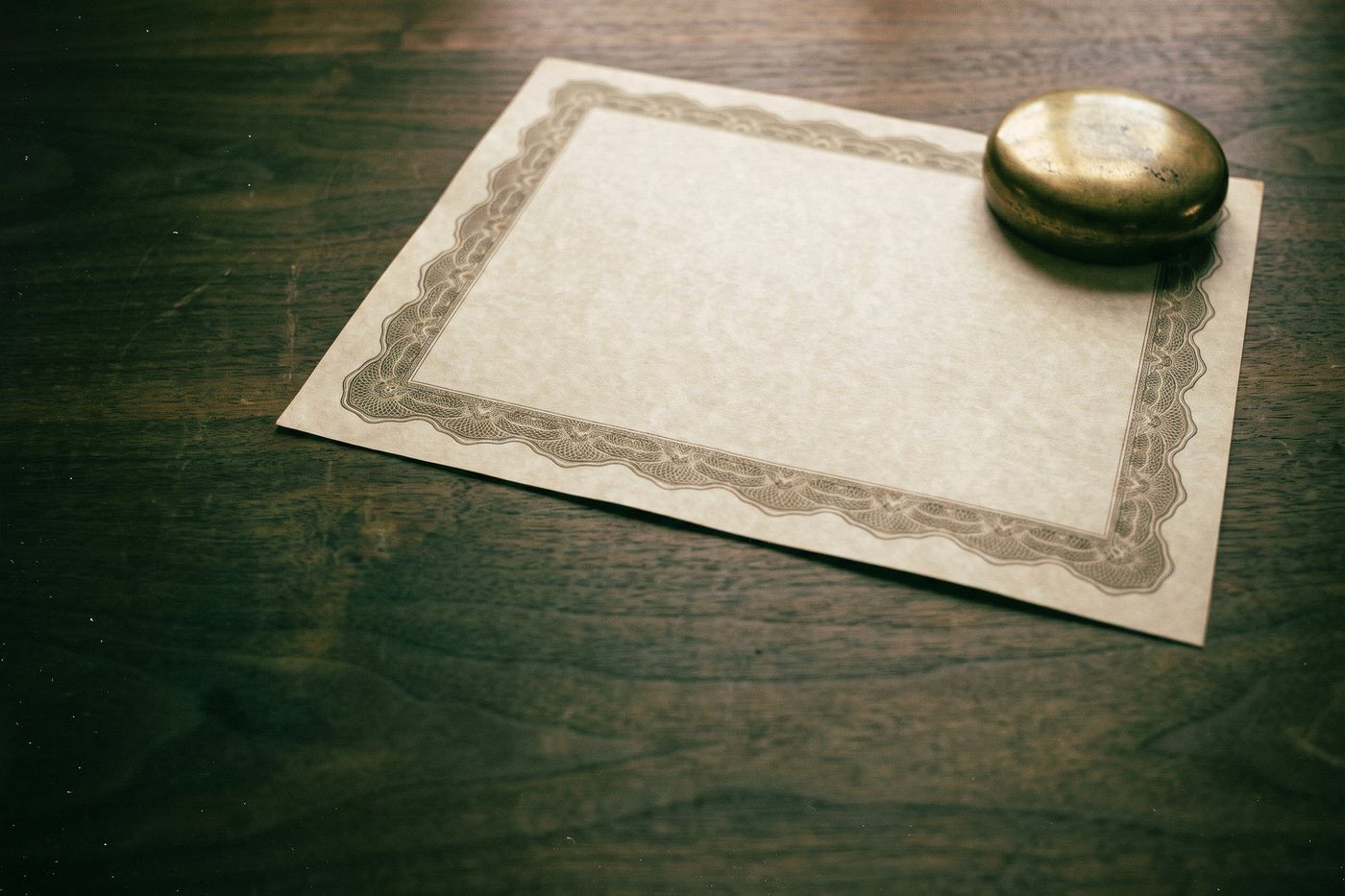 A blank ornate certificate form with an engraved border and no writing, lying on a desk under a brass paperweight