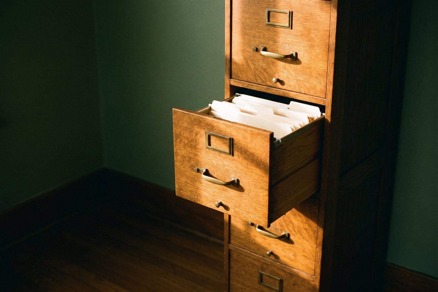 A tall oak filing cabinet with brass drawer pulls, one drawer slightly open showing manila folders with blank tabs
