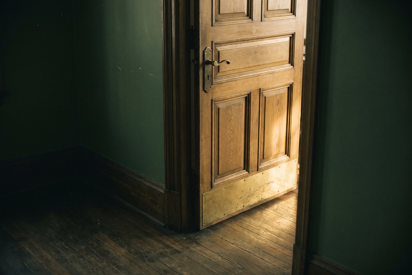 A heavy panelled oak door standing slightly ajar, with a brass lever handle and warm light beyond it