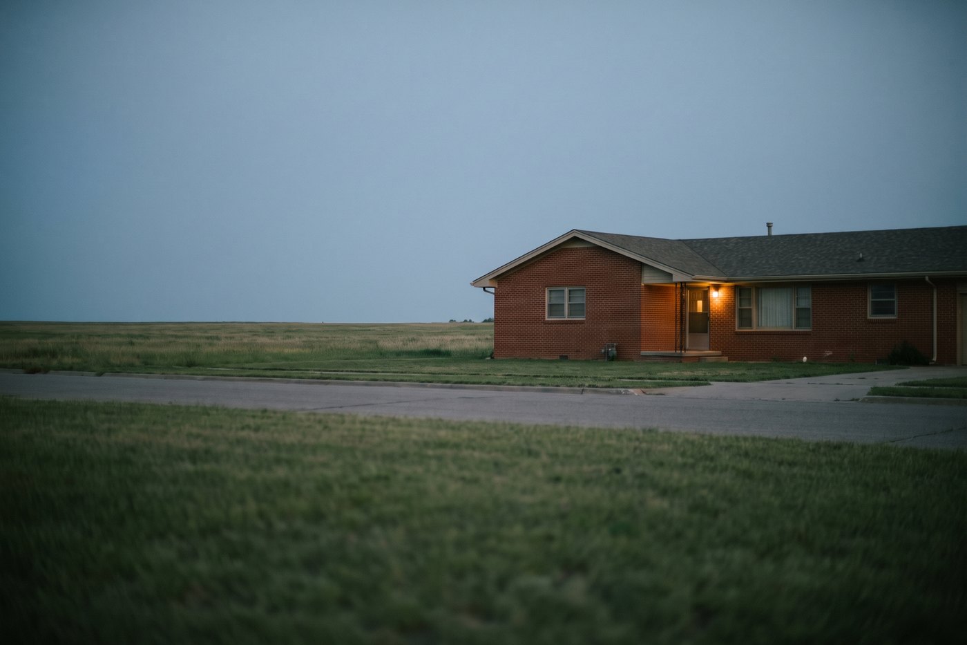 A modest single-storey brick house on a flat grassland street at dusk, a single porch light on