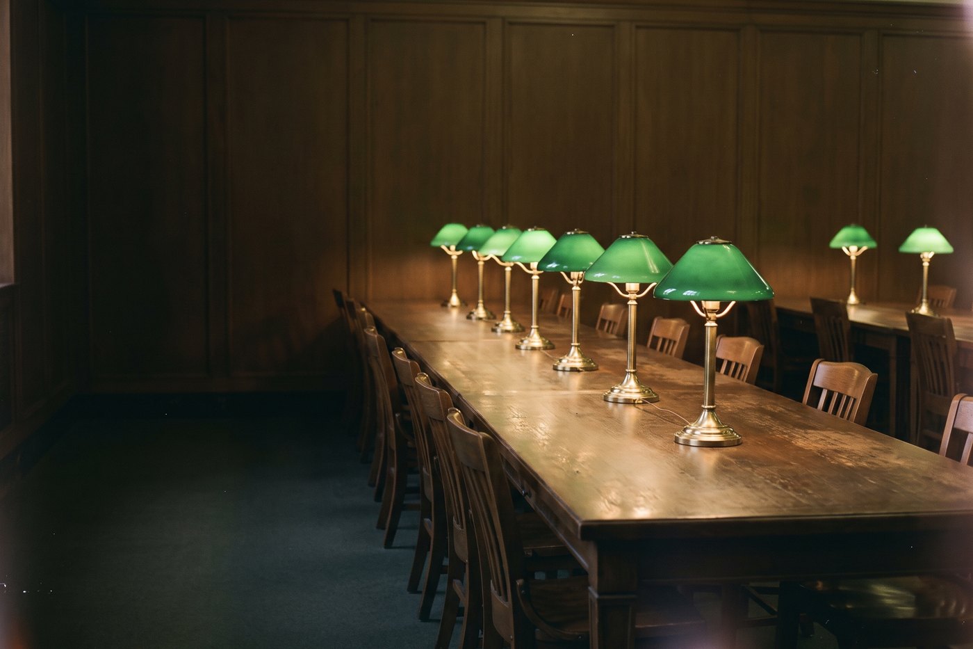 A long empty reading table with a row of green-shaded brass lamps glowing in a dim wood-panelled room
